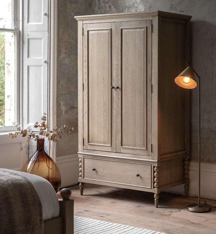 Wooden wardrobe with a drawer, beside a lit floor lamp and a brown vase with dried flowers, set in a rustic room with wooden flooring.