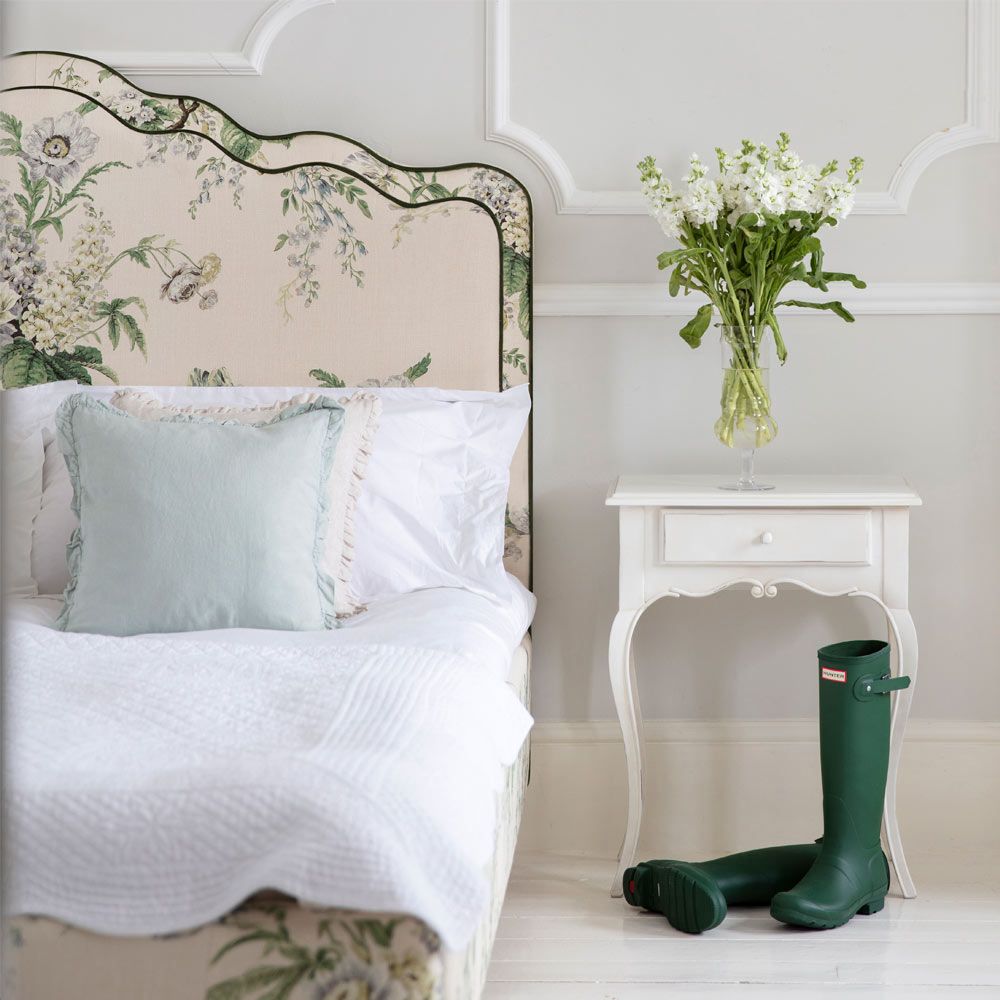 Elegant bedroom with a floral-patterned headboard, white bedding, a side table with a vase of flowers, and green boots on the floor.