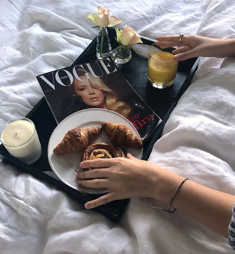 Breakfast in bed with croissants, a magazine, a candle, orange juice, and roses on a tray, with hands reaching for the food.