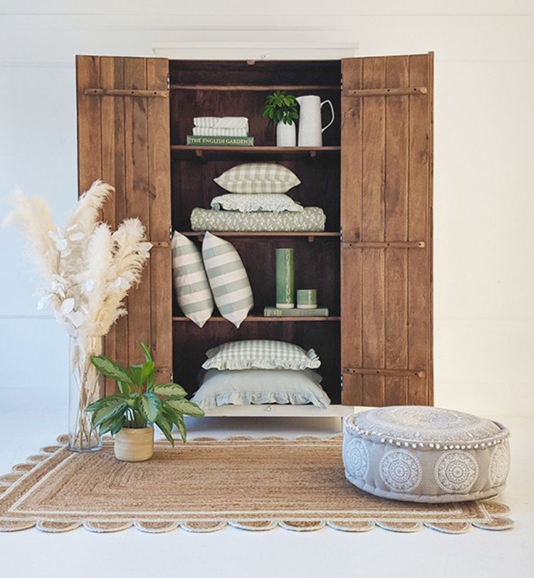 Wooden cabinet with open doors displaying pillows and books, next to pampas grass, a plant, and a patterned pouf on a woven rug.