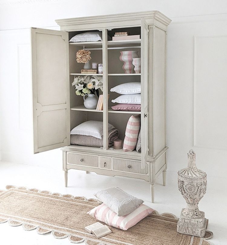 Open vintage cabinet with neatly stacked pastel pillows and blankets, beside a decorative urn and a woven rug on a white floor.