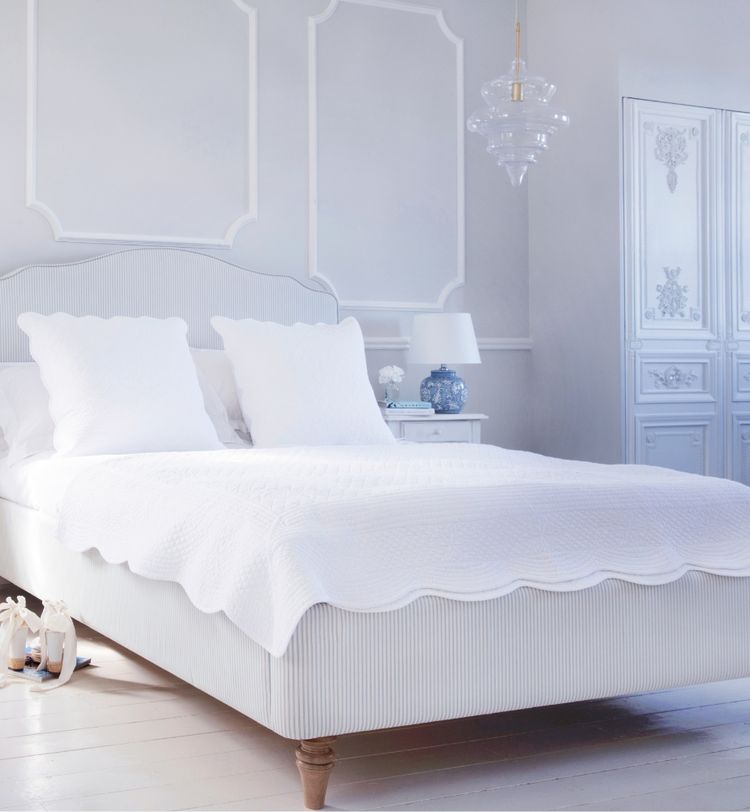 Elegant bedroom with a white bed, textured bedspread, and pillows. Features a bedside table with a lamp and decorative details on the walls and wardrobe.