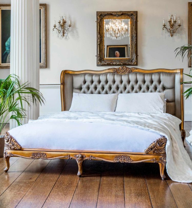 Elegant bedroom with a tufted headboard, ornate wooden bed frame, white bedding, chandeliers, framed art, and potted plants on wooden floor.