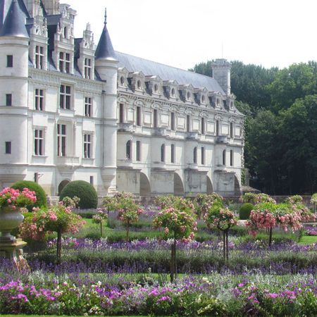 Elegant château with ornate towers and lush gardens, featuring vibrant flowers and manicured lawns under a clear sky.