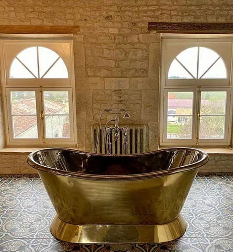A luxurious bronze bathtub sits on patterned tiles between two arched windows in a rustic stone-walled bathroom.