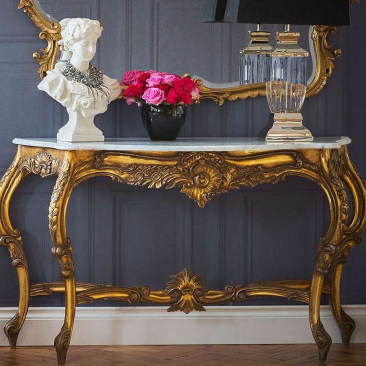 Elegant gold console table with ornate carvings, a marble top, a white bust, a black vase with red roses, and a crystal lamp against a dark wall.