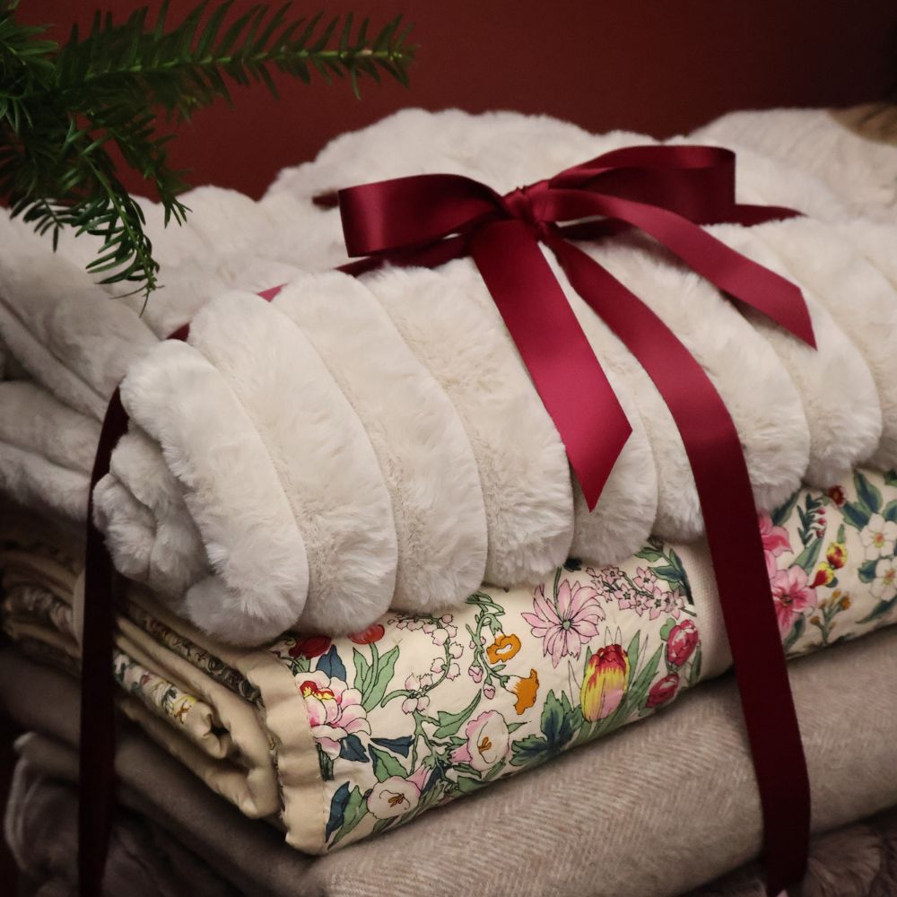 Rolled white faux fur blanket tied with a red ribbon, resting on floral and beige blankets, next to a green plant.