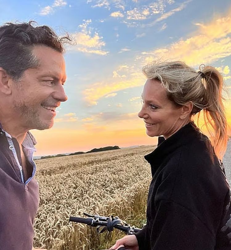 A man and woman smile at each other near a wheat field, with a bicycle and a colorful sunset sky in the background.