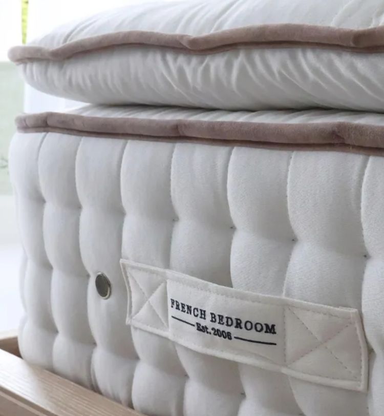 Close-up of a plush, white mattress with a beige trim, featuring a label reading "French Bedroom Est. 2006," placed on a wooden bed frame.