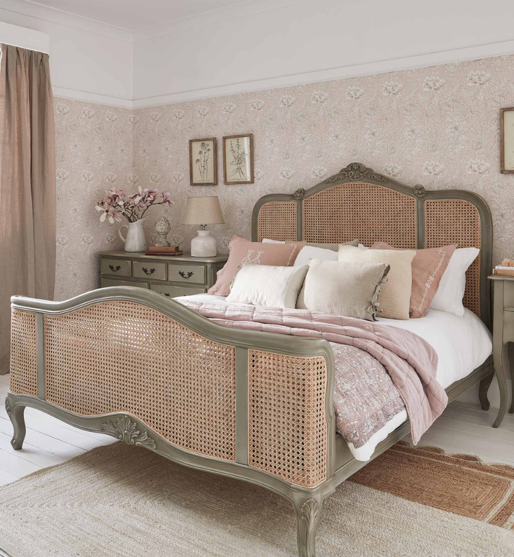 Elegant bedroom with a vintage-style bed, floral wallpaper, soft pink bedding, and a window with brown curtains. Decor includes framed art and flowers.