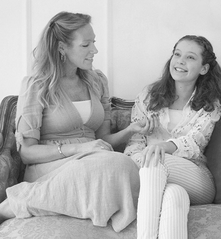 Woman and girl smiling and sitting on a sofa, engaged in conversation. The woman gestures with her hand while the girl looks upward. Black and white.