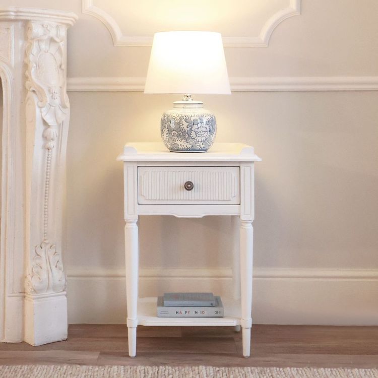 White side table with a drawer, featuring a decorative lamp with a blue floral design. Books are neatly stacked on the lower shelf.