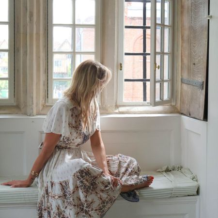 Woman in floral dress sits on a window seat, gazing outside through open windows, with sunlight streaming in.