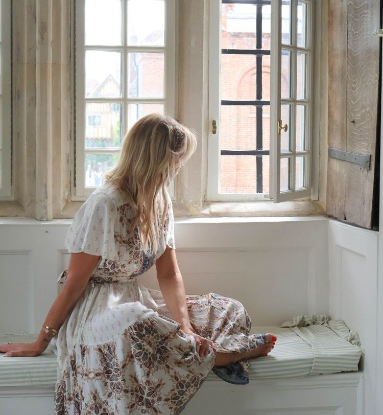 Woman in floral dress sits on a window seat, gazing outside through open windows, with sunlight streaming in.