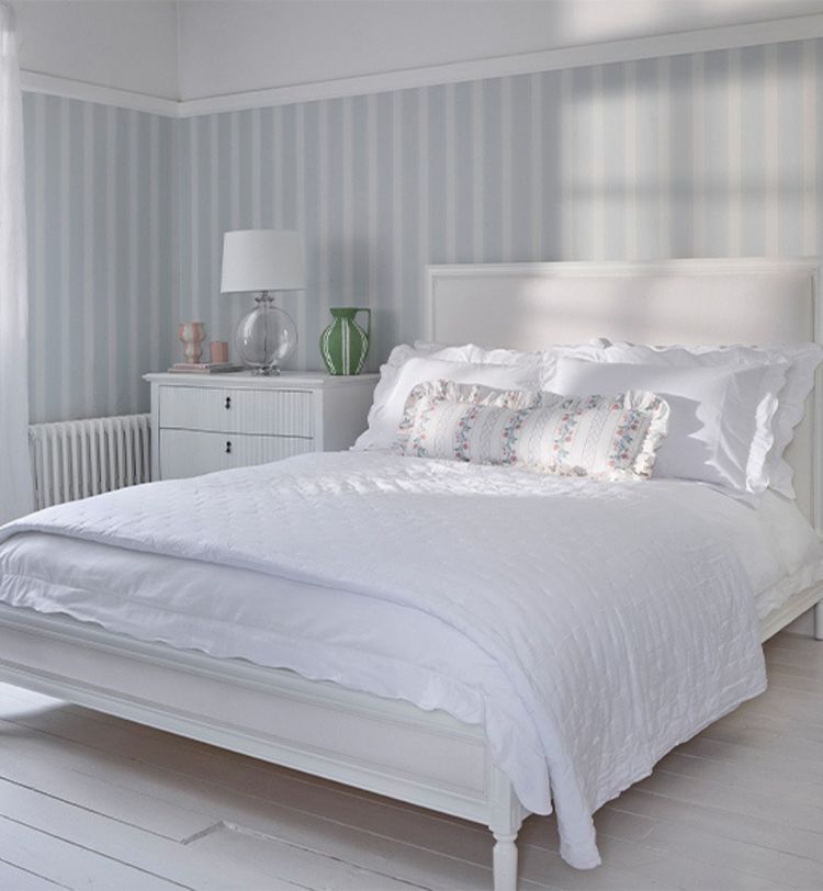 Bright bedroom with a white bed, striped wallpaper, and a side table with a lamp and vases. Soft natural light from a window.