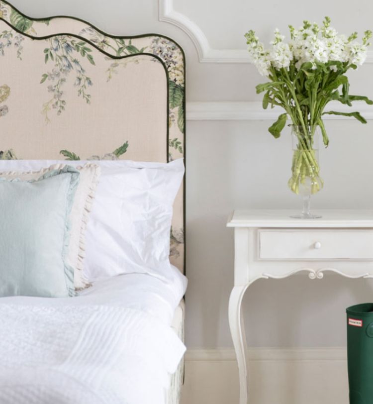 Elegant bedroom with floral headboard, white bedding, and a small white side table holding a vase of flowers. Green boots are on the floor.