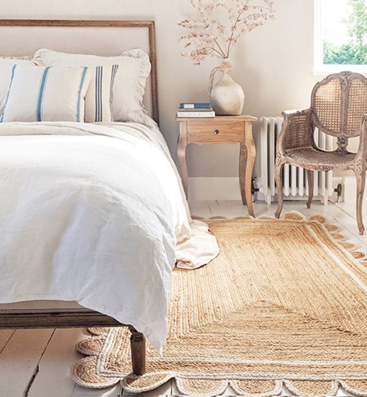 Cozy bedroom with a wooden bed, white linens, a woven rug, a wooden chair, and a small table with a vase of dried flowers.