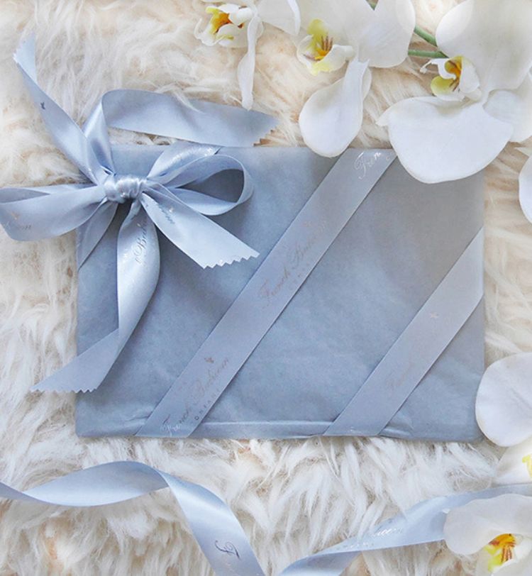 A gift wrapped in light blue paper with matching ribbon, surrounded by white orchids on a fluffy white surface.