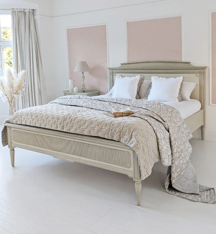 Elegant bedroom with a vintage-style bed, neutral bedding, and soft pink wall panels. A lamp and pampas grass decorate the space.