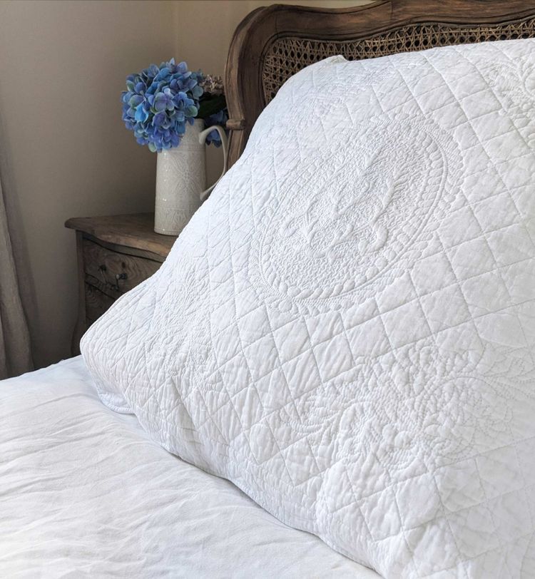 White quilted pillow on a bed with a wooden headboard, next to a nightstand with a white vase holding blue hydrangeas.
