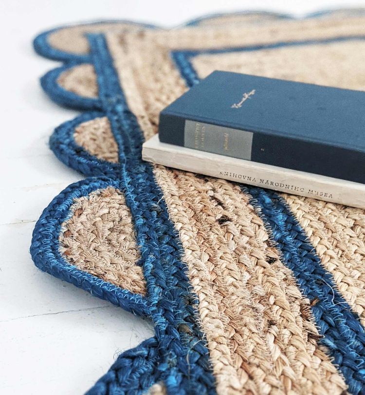 A blue book and a white book placed on a woven jute rug with blue edges and scalloped design, on a white wooden floor.