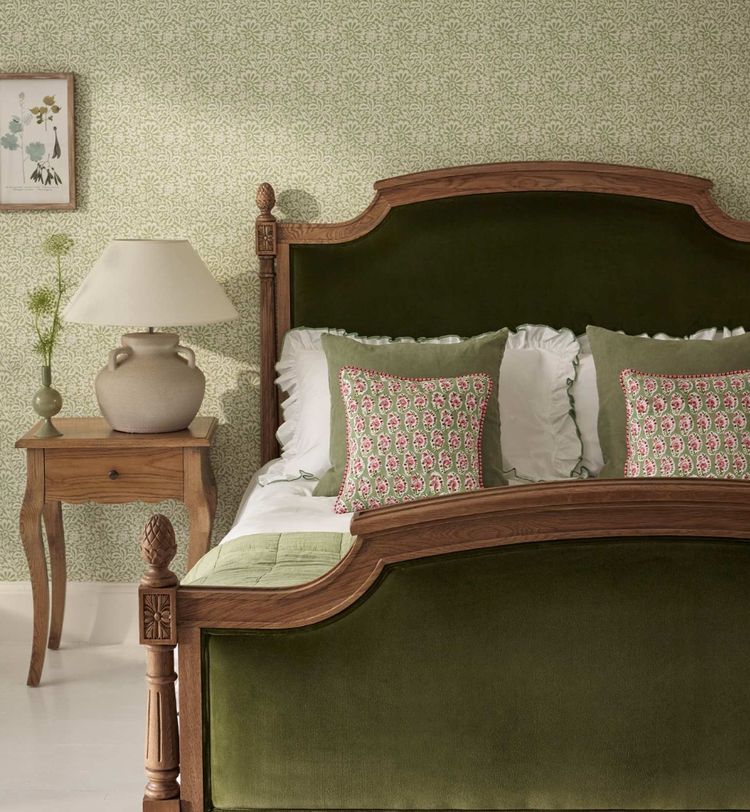Cozy bedroom with a green upholstered bed, floral patterned pillows, a wooden side table with a lamp, and a floral wallpaper backdrop.