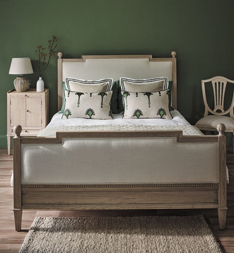 Wooden bed with white and green pillows, beside a nightstand with a lamp and vase. Green wall, wooden chair, and woven rug on the floor.