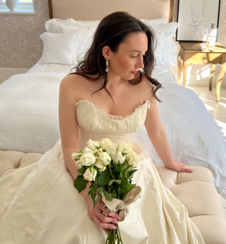 Woman in an elegant white gown holding a bouquet of white roses, sitting on a bed in a softly lit room.