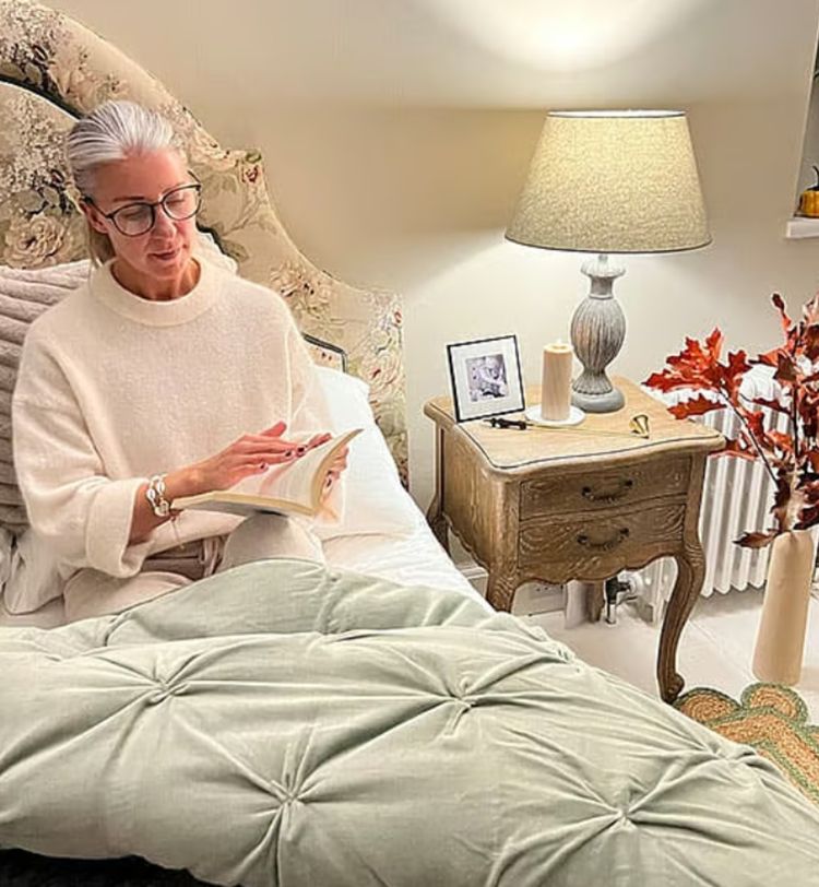 Woman reading in bed with a cozy blanket, beside a lamp-lit nightstand with decor and a vase of autumn leaves.