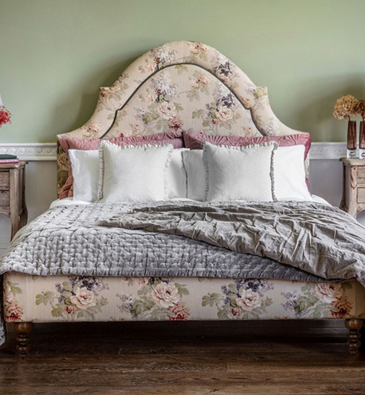 Floral upholstered bed with white and pink pillows, gray quilt, flanked by wooden nightstands with lamps, against a green wall.