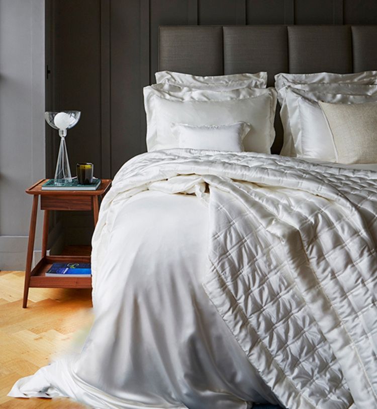 Luxurious bedroom with a neatly made bed featuring white silk sheets and pillows, a wooden nightstand with a lamp, and a dark gray headboard.