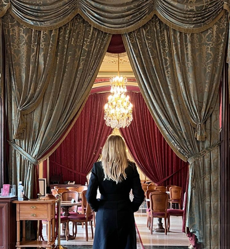 A woman in a black coat stands before ornate drapes, revealing a luxurious room with red curtains and a sparkling chandelier.