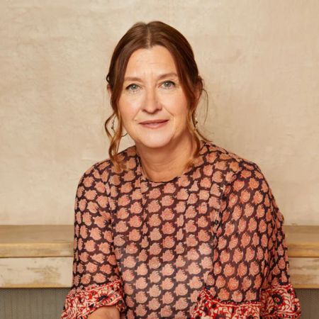Woman with brown hair in a patterned blouse sits against a beige background, smiling softly at the camera.
