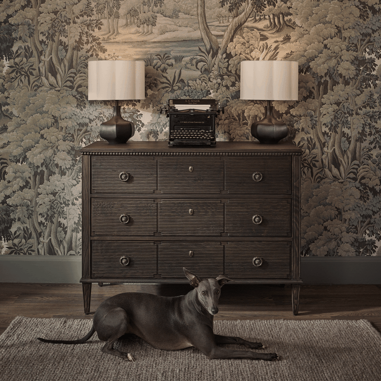 A dog lies on a rug in front of a vintage dresser with a typewriter and lamps, set against a detailed forest-themed wallpaper.