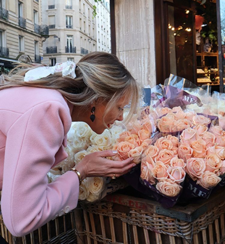 A woman in a pink coat leans over to smell a bouquet of peach-colored roses at a flower stand on a city street.