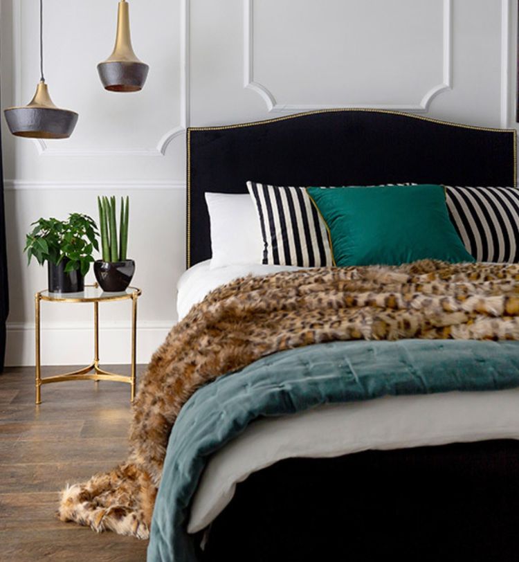 Stylish bedroom with a dark headboard, striped pillows, green and fur throws, pendant lights, and a small side table with plants.