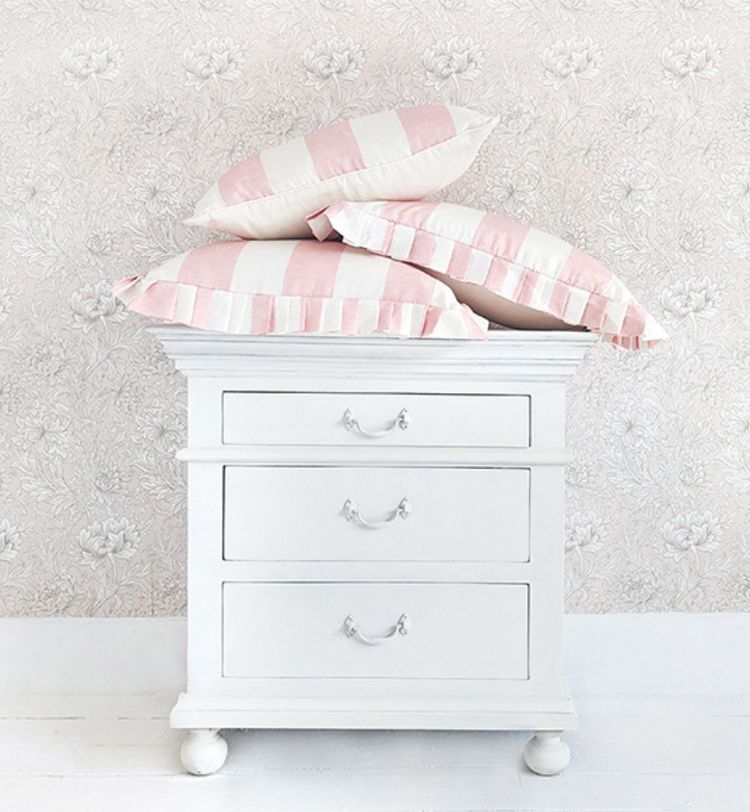 White dresser with three drawers, topped with four pink and white striped pillows, against floral-patterned wallpaper.
