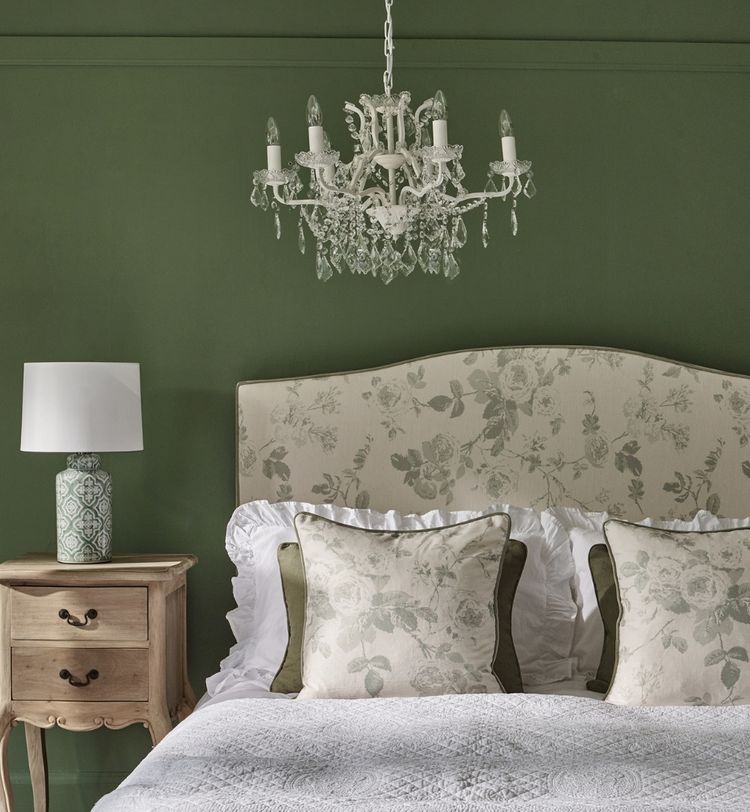 Elegant bedroom with a floral-patterned headboard, two matching pillows, a wooden nightstand with a lamp, and a crystal chandelier overhead.