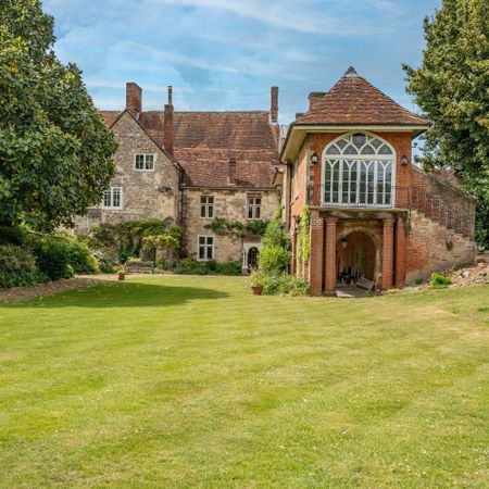 Historic stone manor house with an arched entryway and lush green lawn, surrounded by trees under a blue sky.