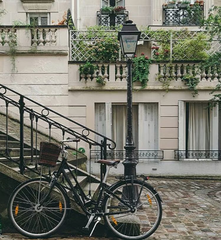 A black bicycle leans against a vintage lamppost near stone steps, with a charming building facade and greenery in the background.