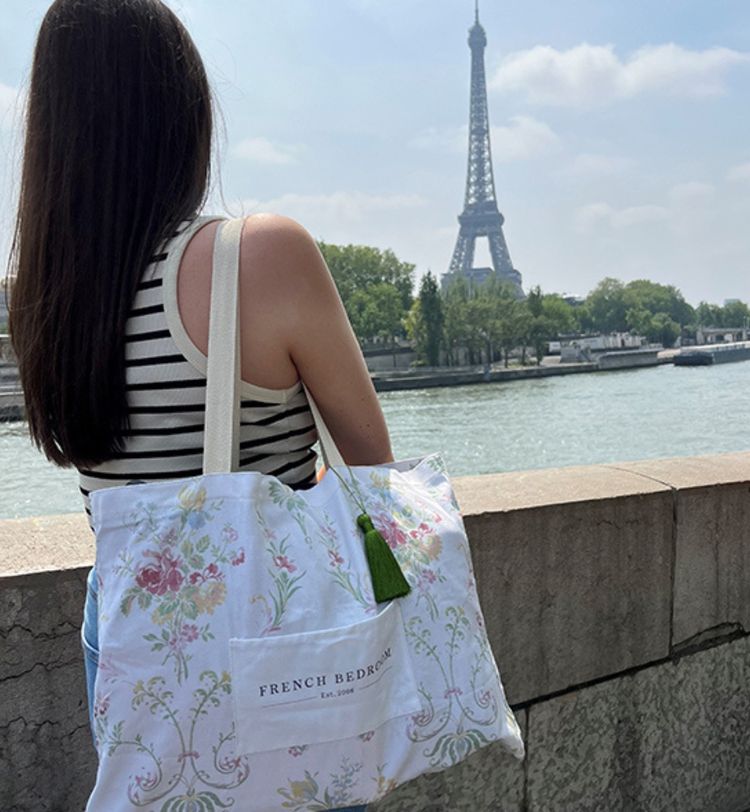 A woman with a striped top and floral bag gazes at the Eiffel Tower by the Seine River on a sunny day.