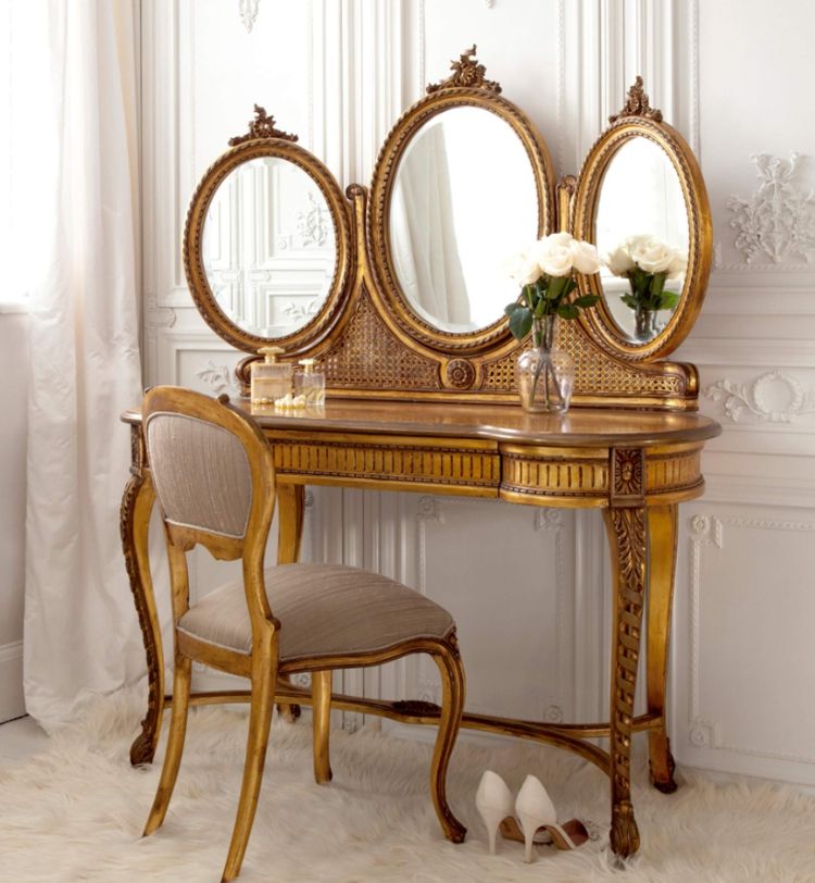 Elegant vanity table with three mirrors, ornate gold detailing, a cushioned chair, white roses, and high heels on a plush rug.