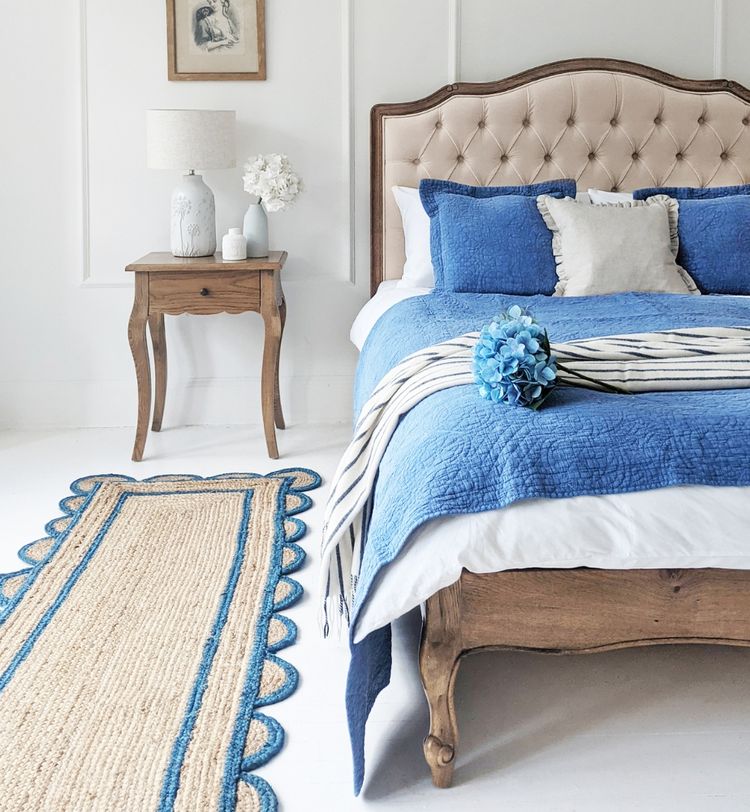 Elegant bedroom with a wooden bed, blue bedding, and a striped throw. A bedside table with a lamp and flowers, plus a woven rug, complete the decor.