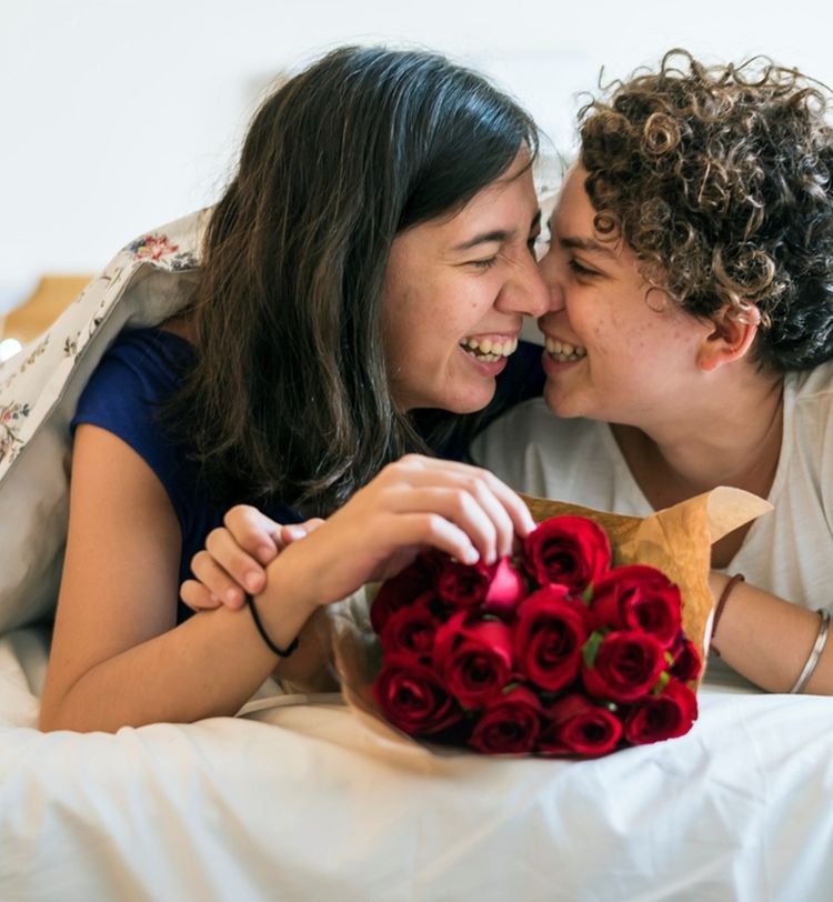 Two people smiling at each other under a blanket, holding a bouquet of red roses, lying on a bed.