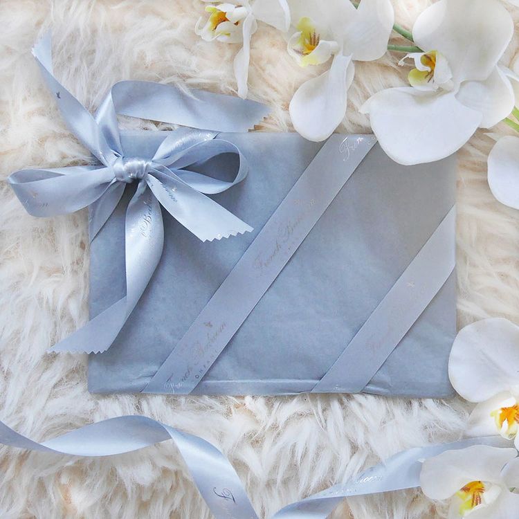 A gift wrapped in light blue paper with matching ribbon, surrounded by white orchids on a fluffy white surface.
