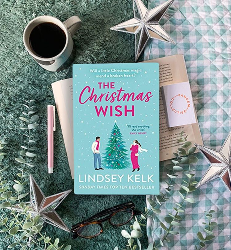 A book titled "The Christmas Wish" by Lindsey Kelk on a cozy setup with a cup of coffee, glasses, and decorative stars on a green blanket.