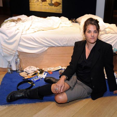 A person sits on a blue rug beside a messy bed with scattered items, including bottles and papers, wearing a black jacket and boots.