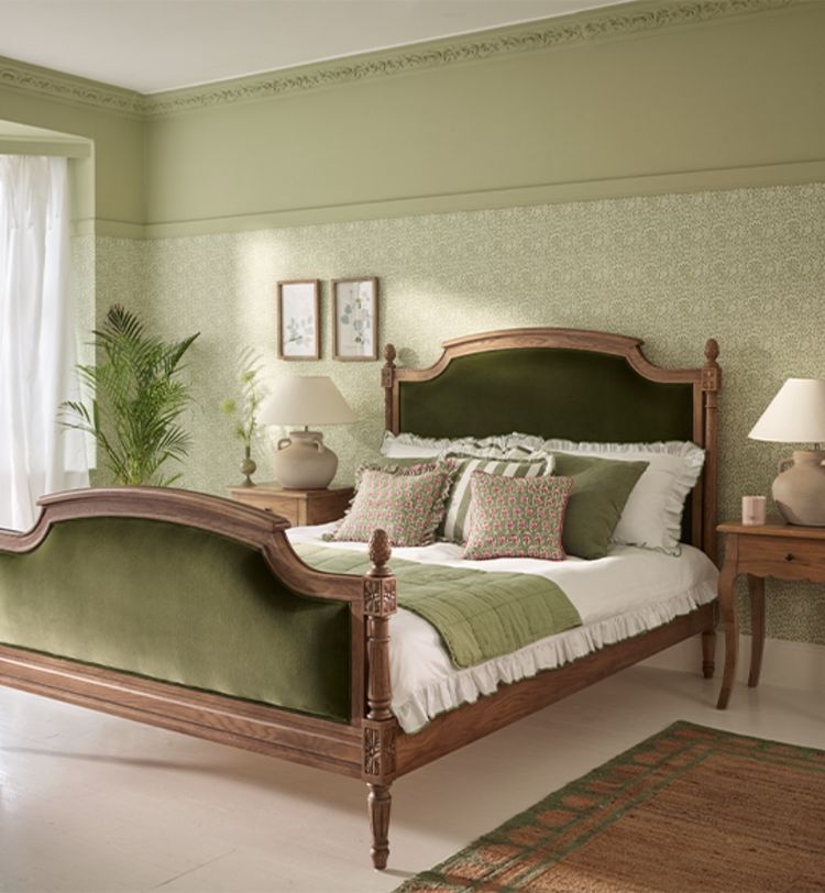 Elegant bedroom with a wooden bed, green velvet accents, patterned wallpaper, and potted plants. Natural light streams through a large window.