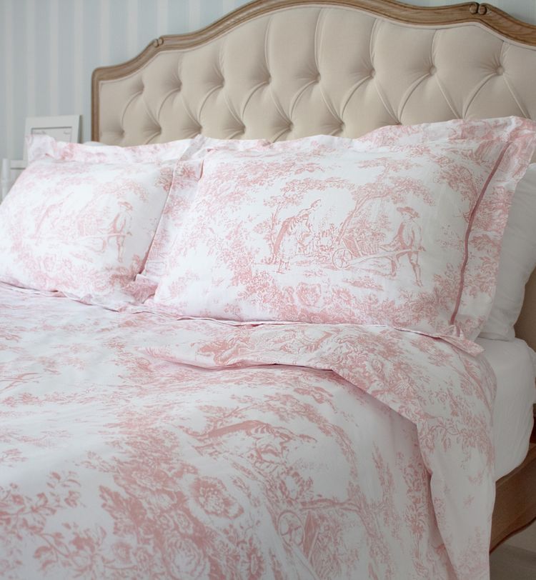 Elegant bed with a tufted headboard, dressed in pink and white toile-patterned bedding, in a softly lit bedroom.