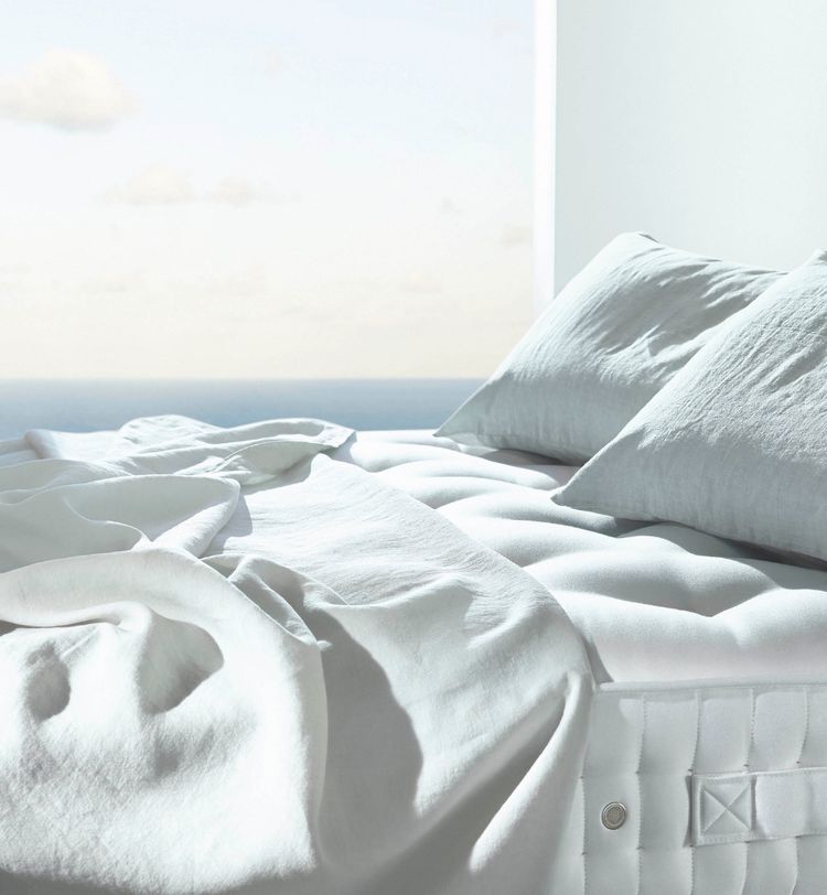 A neatly made bed with white linens and pillows, set against a bright window with an ocean view in the background.